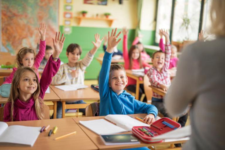 Children in a classroom, holding their hands up to answer a question